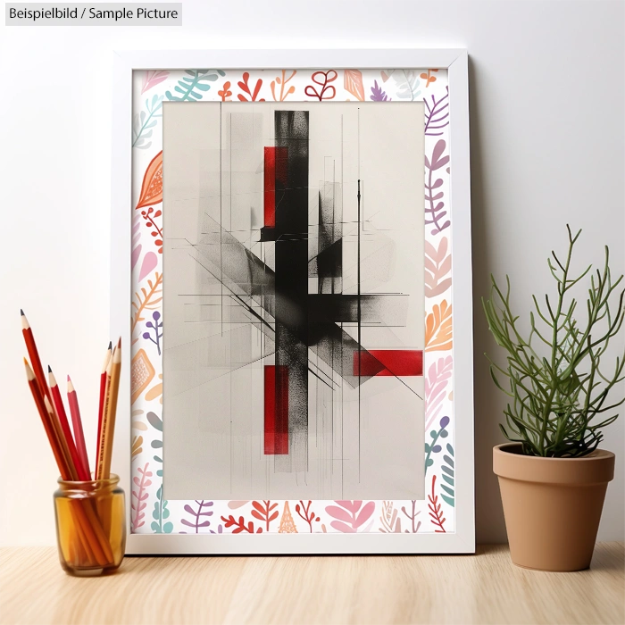 Abstract artwork with black and red geometric lines, framed with floral design, next to a potted plant and pencils.