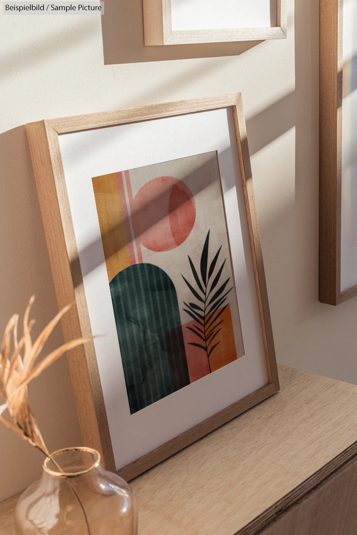 Framed abstract art with geometric shapes and a plant, leaning on a wooden surface in sunlight.