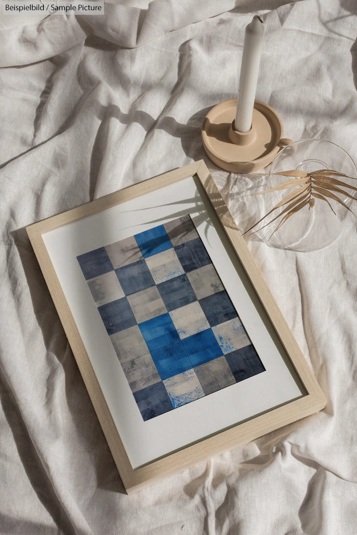 Framed abstract blue checkered artwork on crumpled fabric with candle and glass vase.