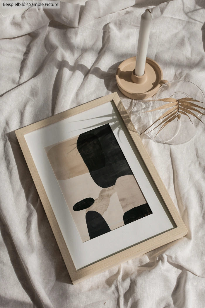 Framed abstract art with black, beige shapes on white linens, beside glass vase and candle on wooden holder.