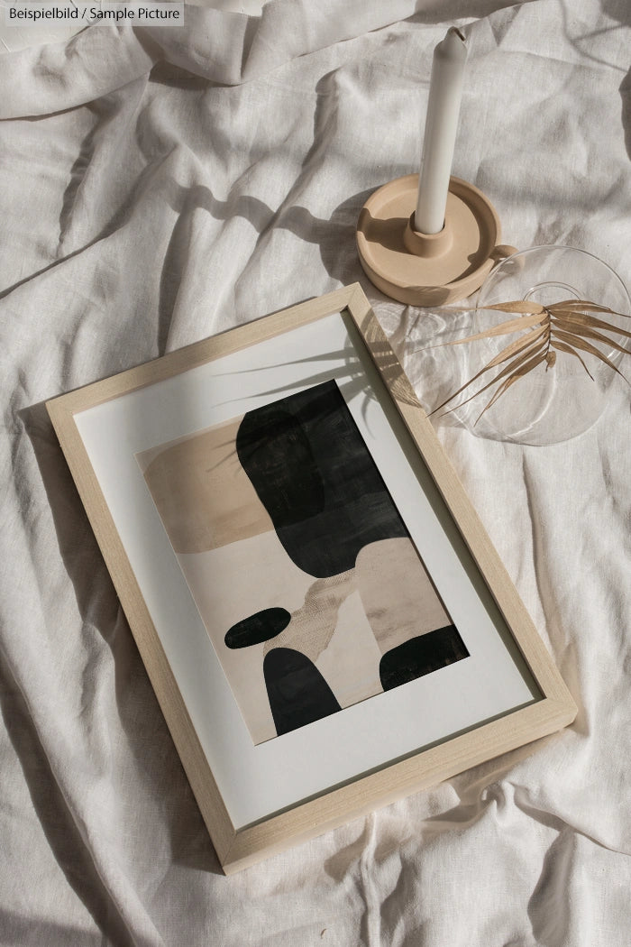 Framed abstract art with black, beige shapes on white linens, beside glass vase and candle on wooden holder.