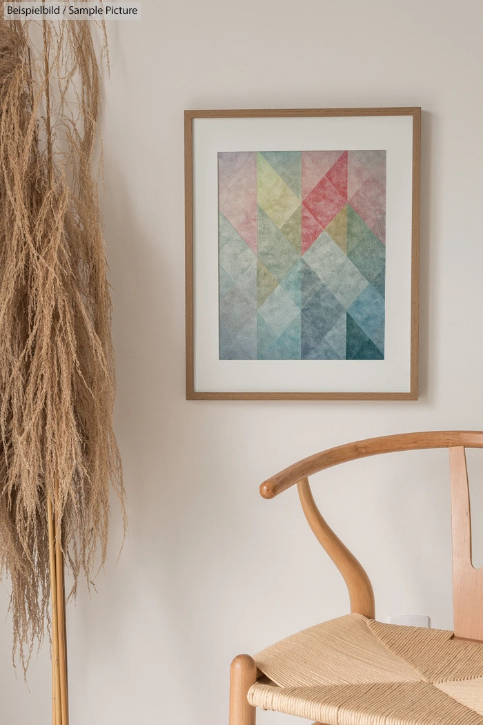 Geometric abstract art in pastel colors framed on a cream wall beside a wooden chair and dried pampas grass.