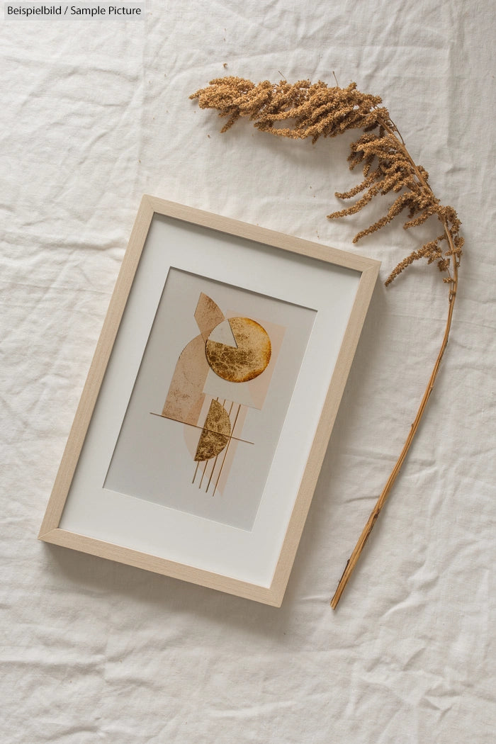 Framed abstract art with geometric shapes on white fabric and dried plant stem on the side.