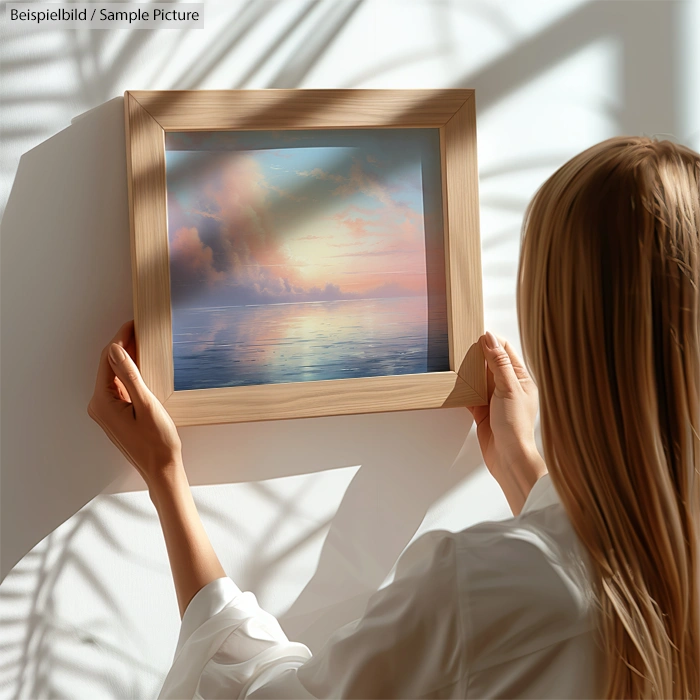 Person hanging framed sunset seascape painting on a white wall.