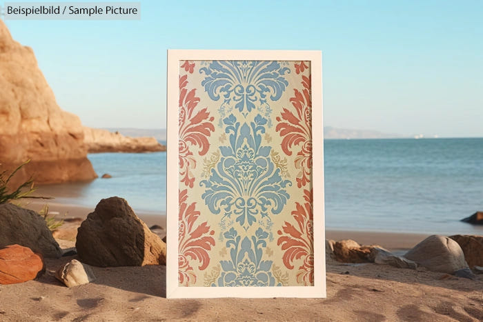 Intricate patterned artwork in frame displayed on rocky beach with ocean view.