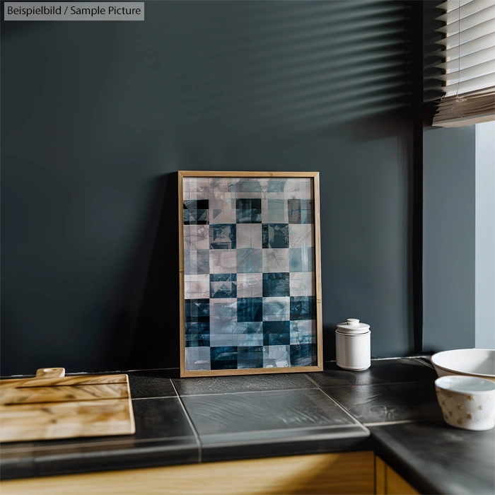 Framed abstract geometric artwork with blue and gray squares on a dark kitchen wall beside a wooden counter.