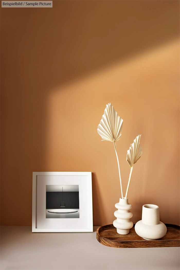 Minimalist decor with a framed photo, ceramic vases, and dry palm leaves on a wooden tray against a beige wall.