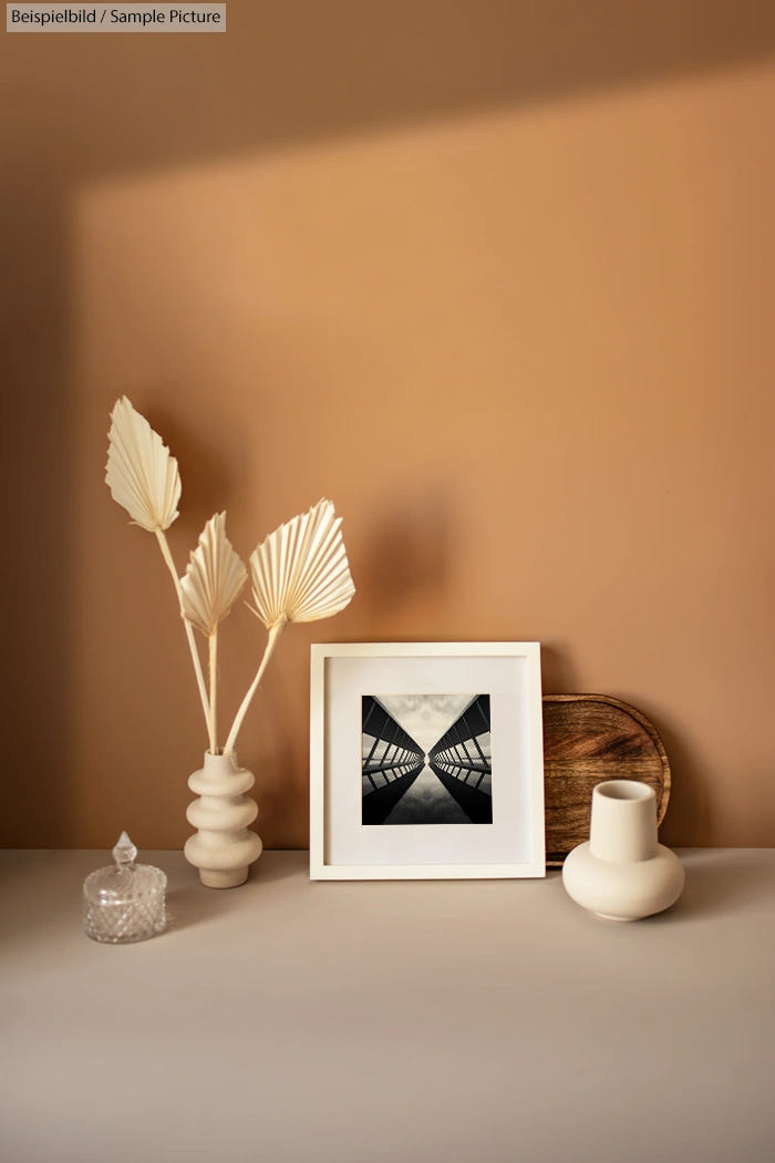 Minimalist shelf display with a framed geometric photo, ceramic vases, and decorative leaf arrangement against a beige wall.