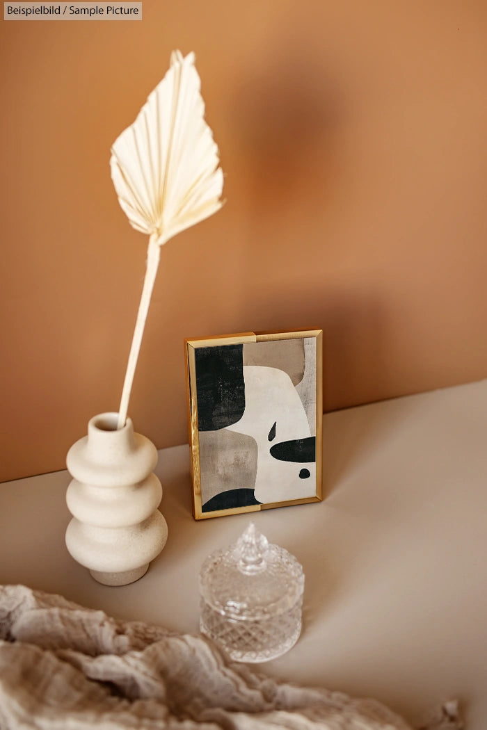 Minimalist decor with abstract art, ceramic vase with sculptural leaf, and glass jar on beige surface.