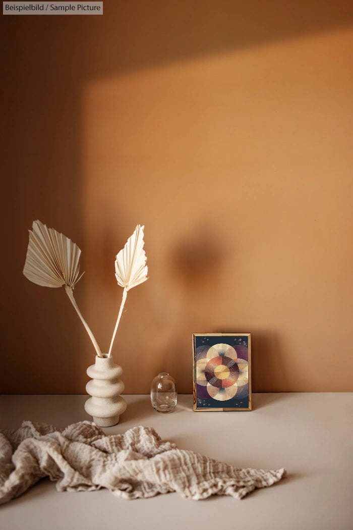 Minimalist vase with dried leaves, abstract framed artwork on a beige wall, fabric draped on a white surface.