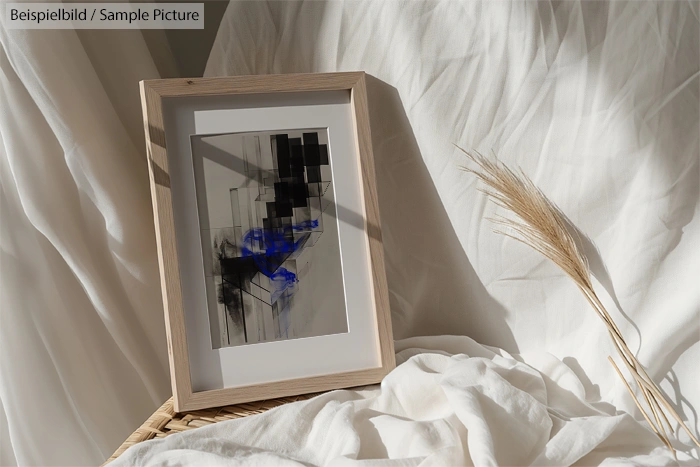 Framed abstract artwork with geometric shapes in black and blue, set against a white fabric background with dried flowers.