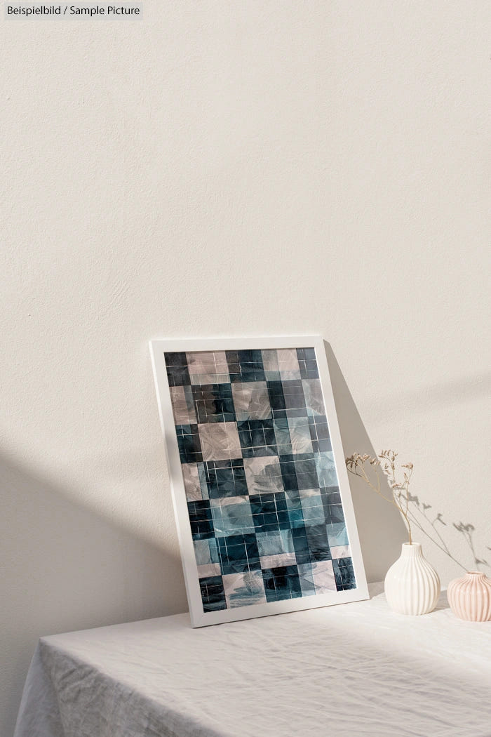 Decorative framed artwork with a grid of abstract patterns, leaning against a white wall on a table with dried flowers.