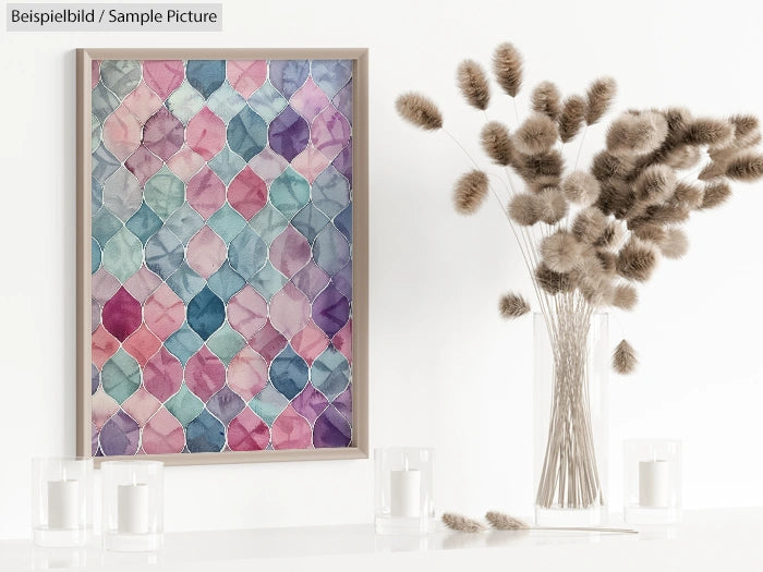 Framed abstract pastel painting with geometric pattern next to vase of dried flowers and candles on a white surface.