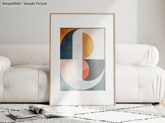 Framed geometric abstract art on floor in front of white couch, with open book on a patterned rug.