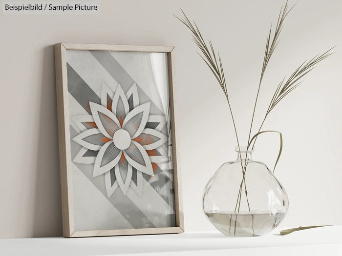 Framed geometric art with floral design beside a glass vase with water and dried reeds on a white shelf.