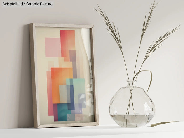 Framed abstract geometric artwork with pastel colors beside a clear glass vase holding tall grasses on a white shelf.