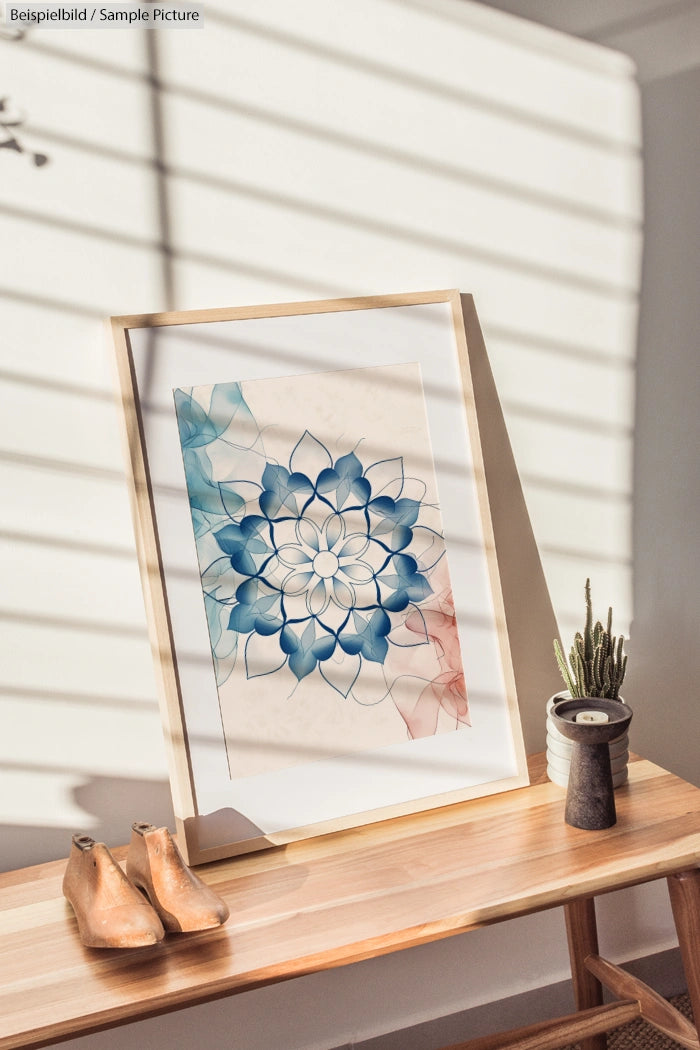 Framed geometric artwork with blue and red floral design on wooden table, sunlight streaming in through blinds.