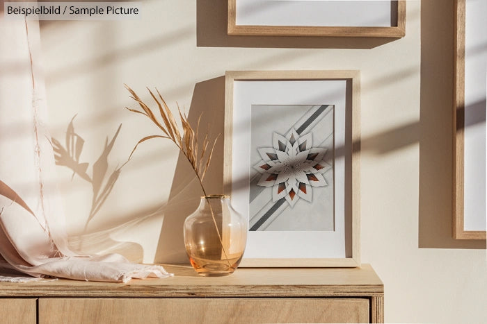 Framed geometric art on a wooden console with a glass vase and dried plant in a sunlit room.