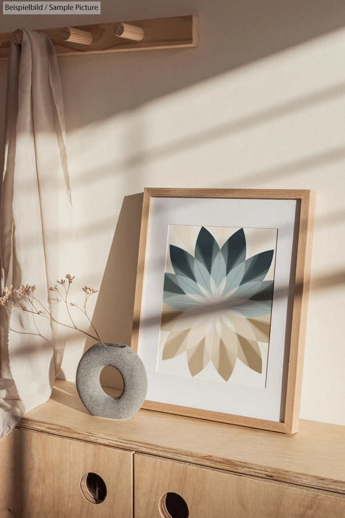 Framed geometric leaf artwork on wooden shelf with round vase, soft shadows and draped fabric in gentle daylight.