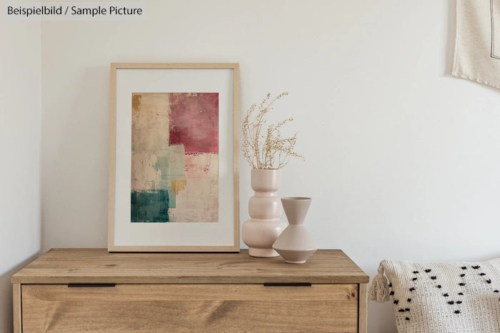 Framed abstract art on wooden dresser with ceramic vases and dried flowers in minimalist room.