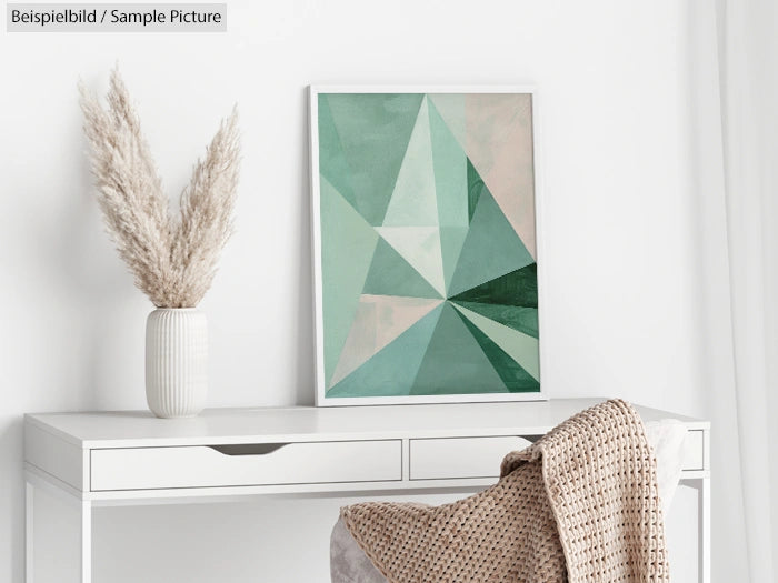 Geometric painting with green and beige triangles on a desk with a white vase and dried grass.