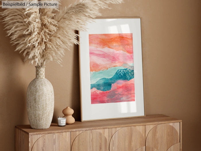 Framed abstract painting with warm tones on a wooden cabinet, next to pampas grass in a decorative vase.