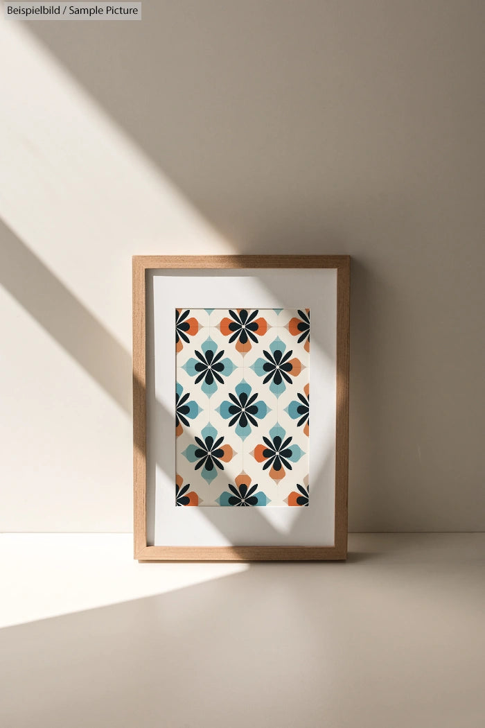 Framed artwork with geometric floral pattern in teal, orange, and blue, leaning against a light beige wall in soft sunlight.