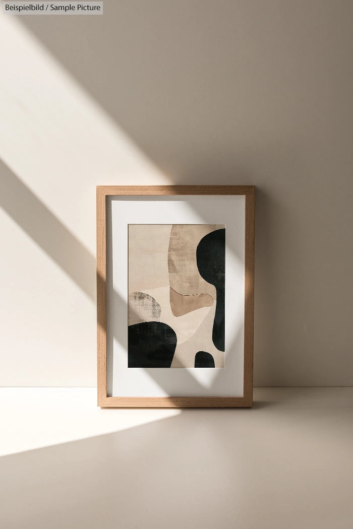 Framed abstract art with beige and black shapes in sunlight against a neutral wall.