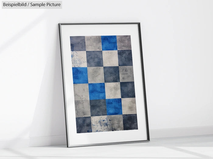 Framed abstract artwork with a grid of blue, gray, and white textured squares leaning against a white wall.