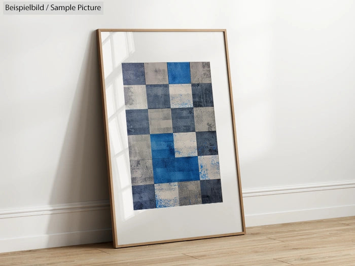 Framed abstract artwork with blue and gray squares leaning against a white wall on wooden floor.