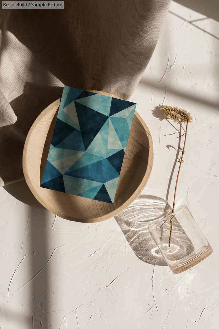 Geometric blue card on wooden plate, glass vase with dried flower on white textured surface, sunlight casting shadows.