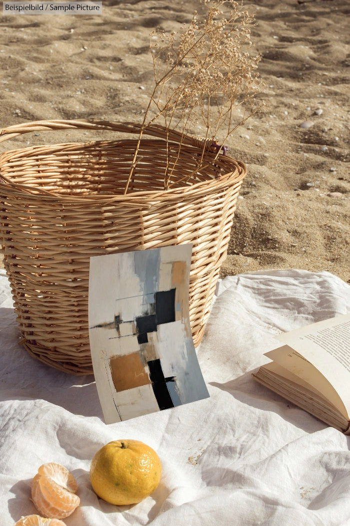 Wicker basket on sandy beach with abstract painting, open book, orange, and lemon on white cloth.