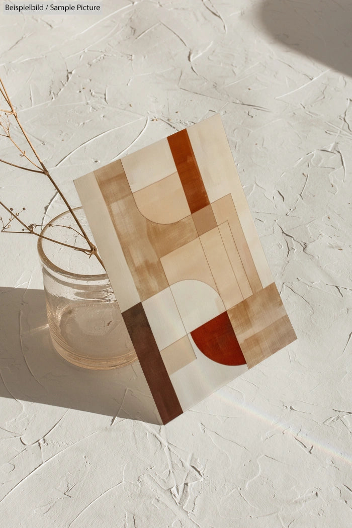 Abstract geometric art print with brown, beige, and red shapes on textured surface next to glass jar with dried twig.