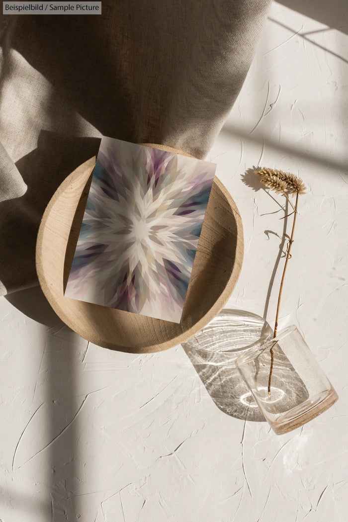 Abstract geometric artwork in wooden bowl on textured surface with dried flower in glass vase, sunlight casting shadows.