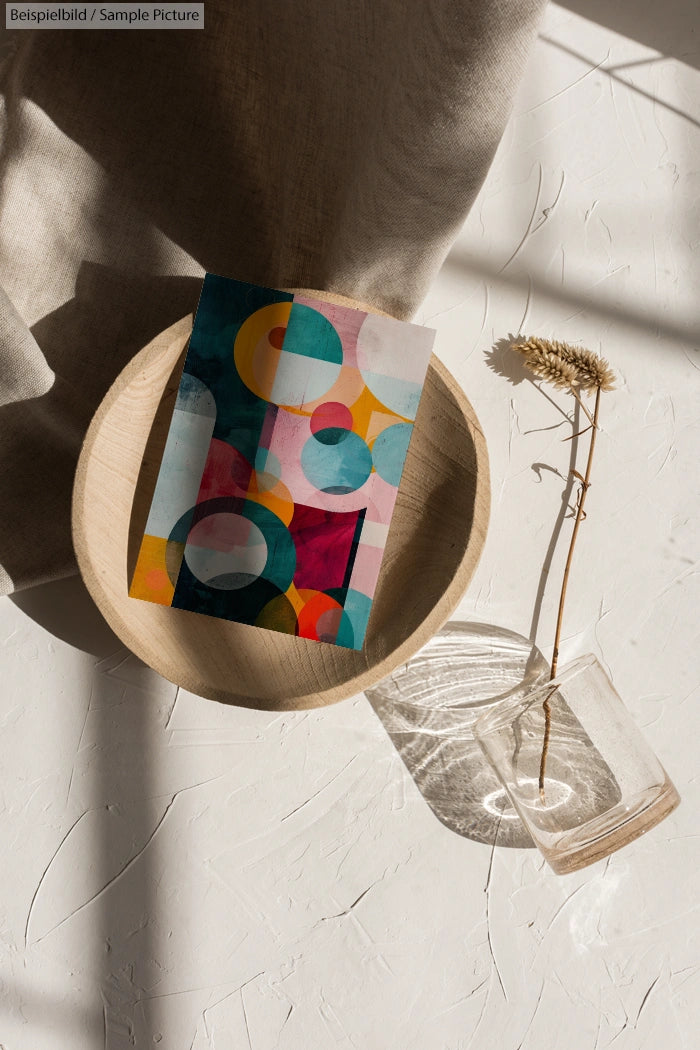 Colorful abstract art piece in round wooden bowl on textured white surface, with a dried flower in a glass vase nearby.