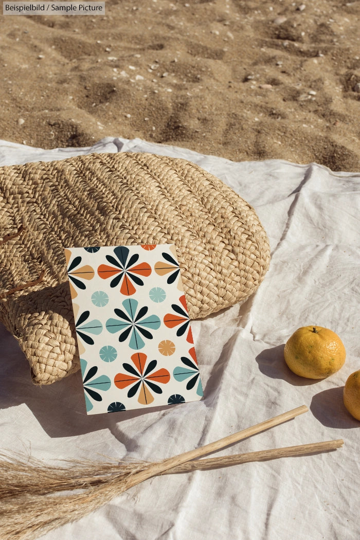 Colorful geometric patterned card on a white cloth with two oranges and straw mat on a sandy beach.