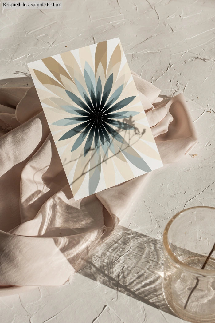 Decorative card with radial flower pattern in neutral tones on textured surface with draped fabric and glass cup nearby.