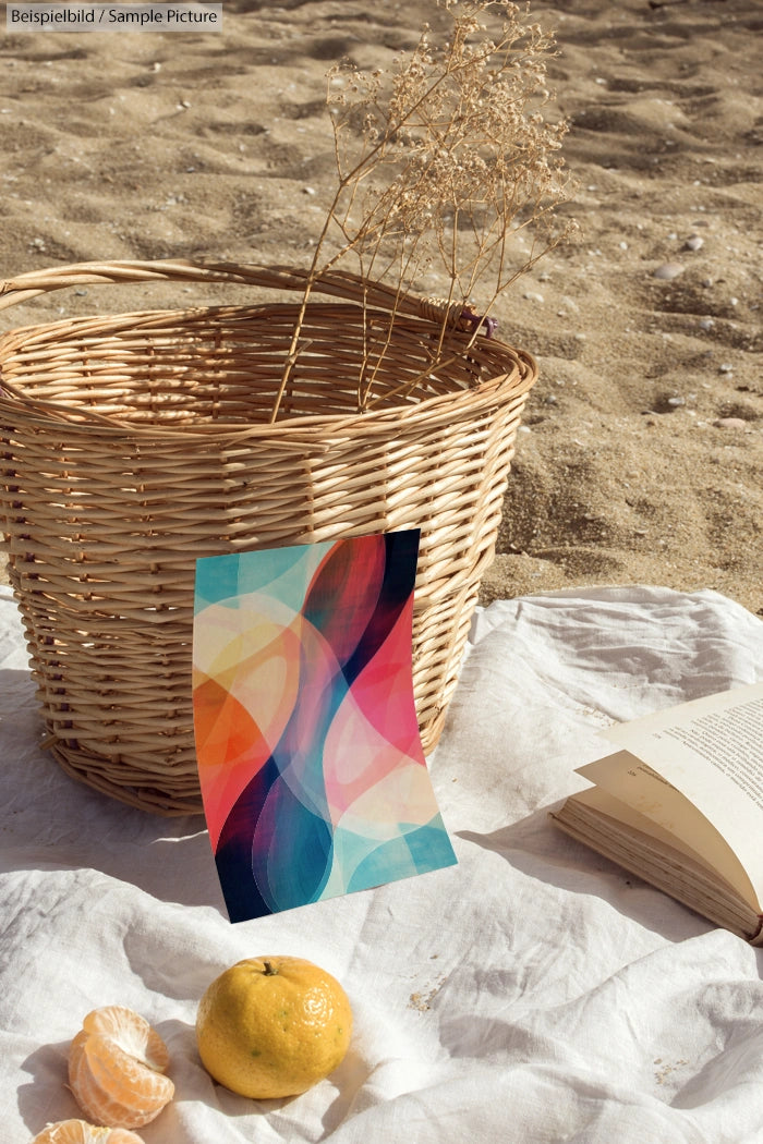 Colorful abstract art near wicker basket, book, and fruits on sandy beach setting.