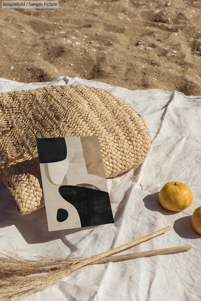 Beach scene with abstract art, woven pillow, oranges, and decorative grasses on sand and white fabric.