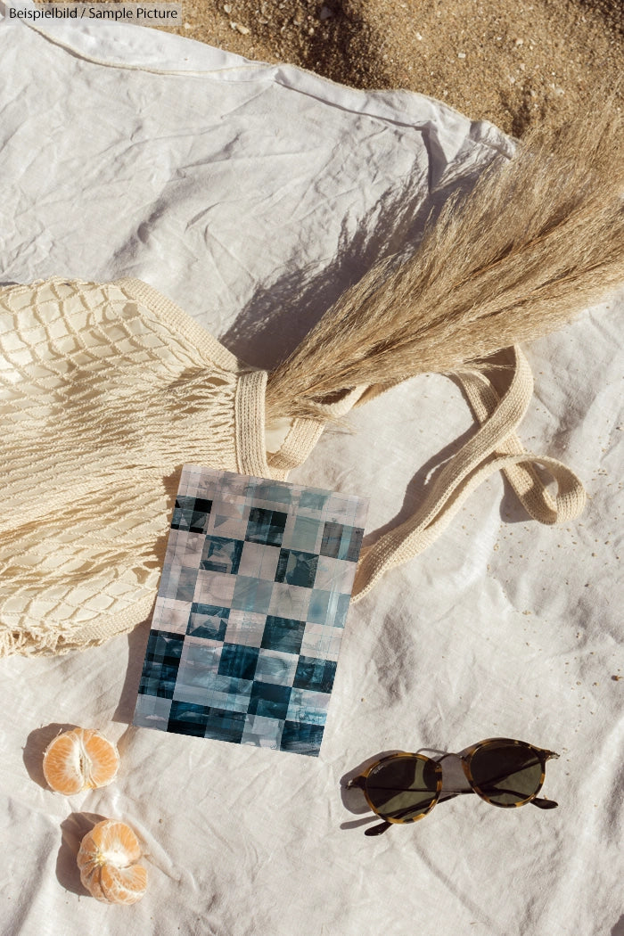 Beach picnic setup with bag, pampas grass, sunglasses, a card, and peeled oranges on a white cloth.