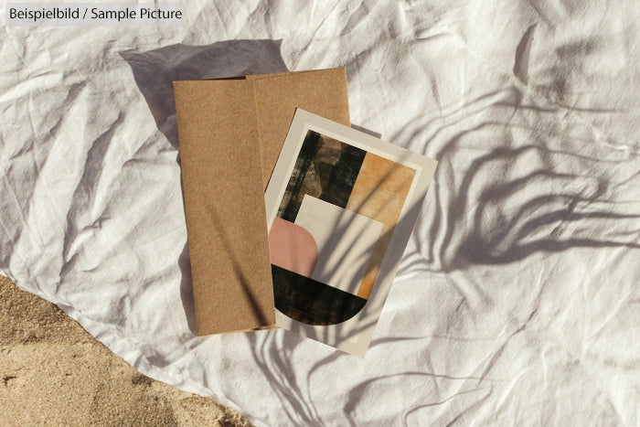 Abstract art print on a sandy beach with brown envelope and textile backdrop.