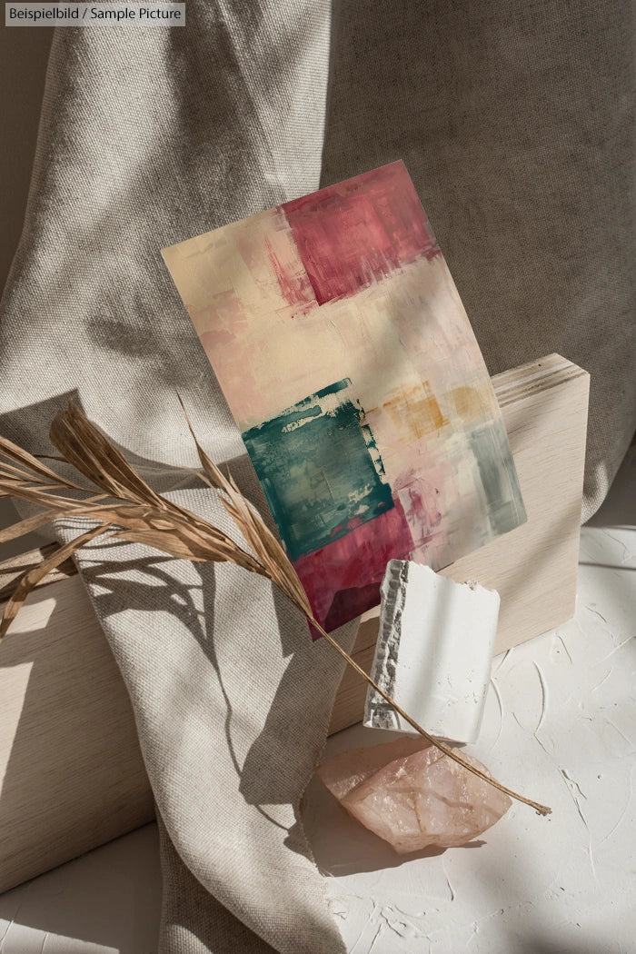 Abstract art with red, teal, and beige hues displayed on textured fabric with dried plant and stone decor.