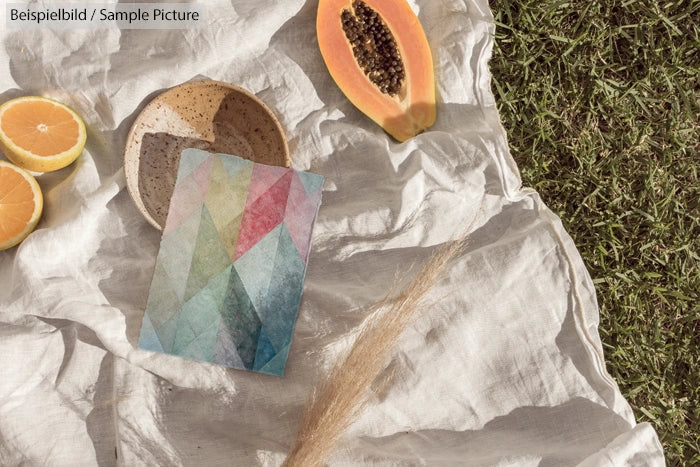 Colorful card on linen with oranges, papaya, and bowl; placed on grass. Bright and artistic outdoor setup.