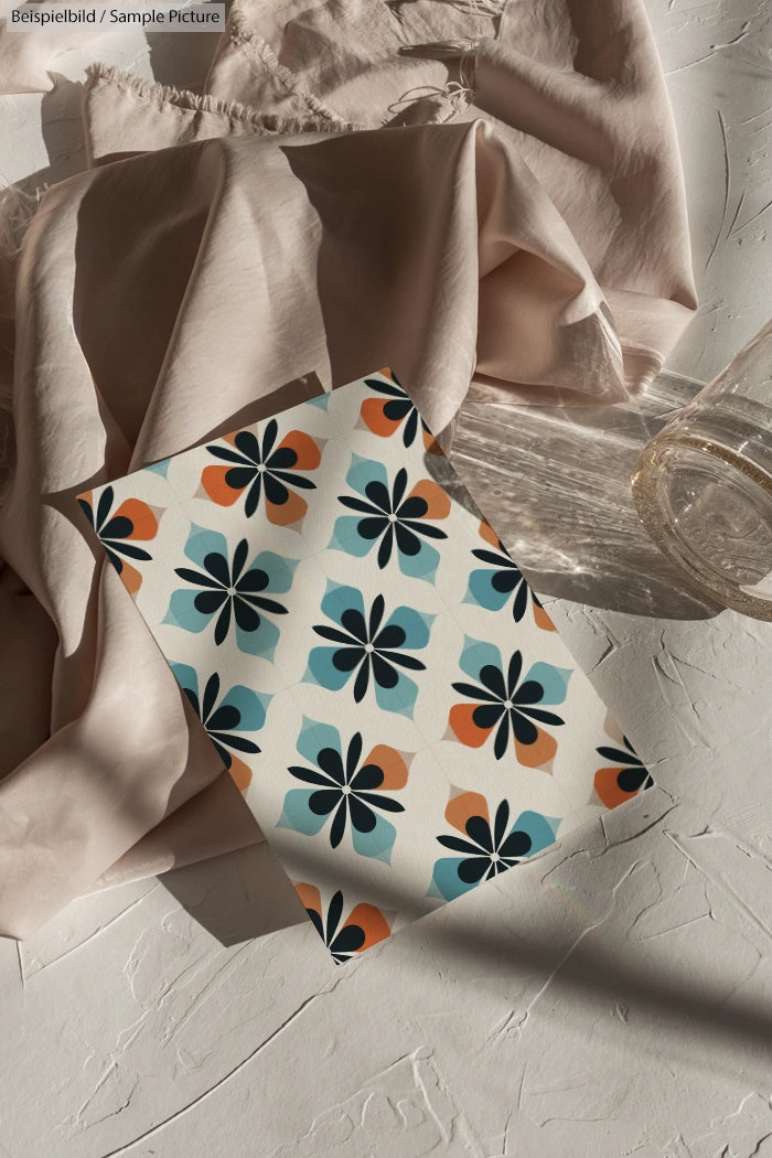 Patterned paper with floral shapes in blue, orange, and black on a textured surface next to beige fabric and a glass.