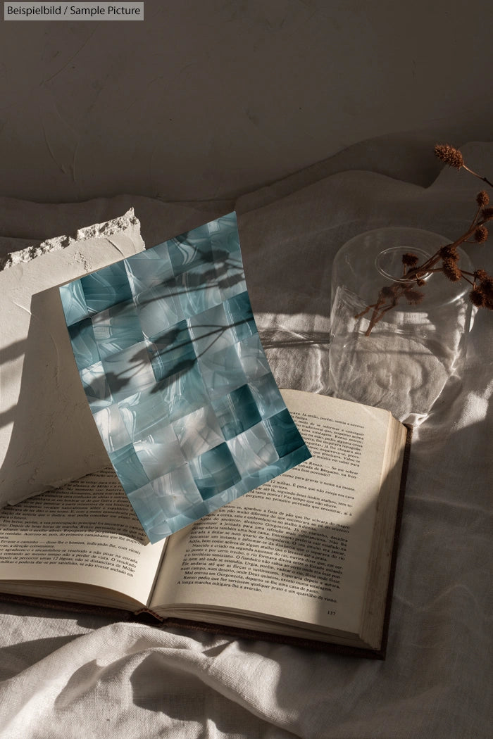 Open book on linen with abstract blue and gray painting, dried flower, and sunlight creating shadows.