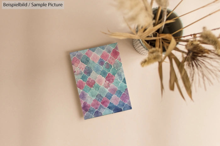 Decorative card with colorful geometric pattern displayed on beige wall with a vase of dried flowers.