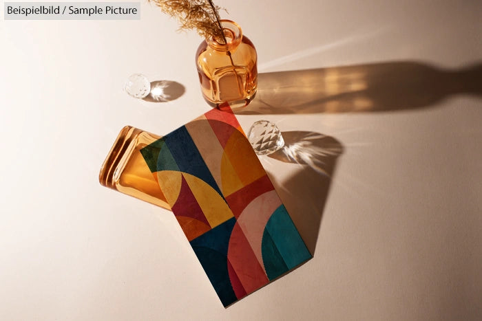 Colorful geometric pattern card beside amber glass bottle on beige surface with plant and crystals.