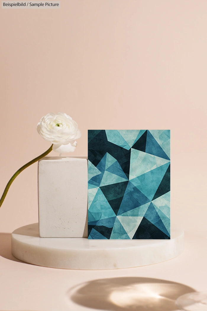 Geometric abstract art with blue shades on easel beside a white flower in a minimalist setting.