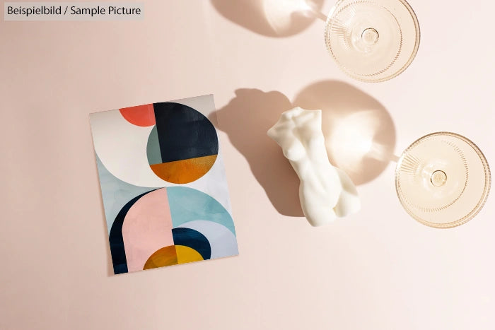 Abstract geometric art beside a white sculpture and two glass cups on a beige surface.