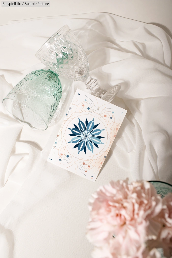 Elegant card with blue floral design beside crystal glasses on draped white fabric, pink flower in foreground.